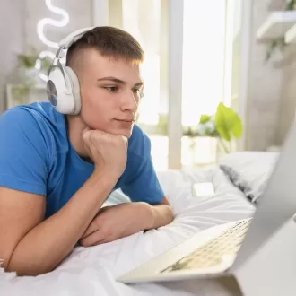 A teenage boy wearing headphones looking at a laptop on his bed