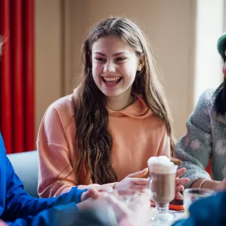 A group of happy looking teenagers sat around a dining table drinking hot chocolate and smiling