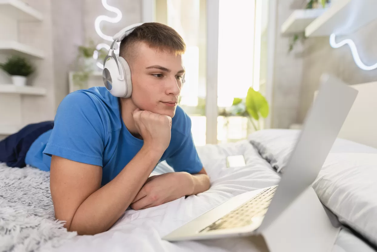 A teenage boy wearing headphones looking at a laptop on his bed