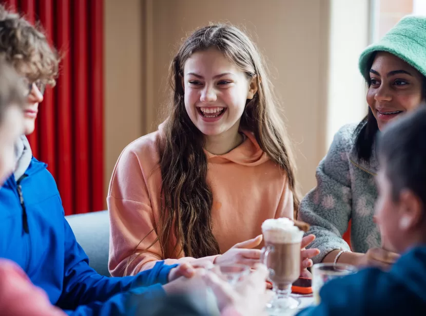 A group of happy looking teenagers sat around a dining table drinking hot chocolate and smiling